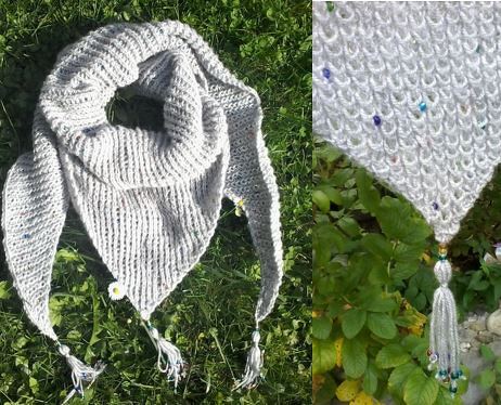 Graues gestricktes Dreieckstuch mit bunten Perlen, Quasten und einer kleinen weißen Blüte auf grünem Gras.