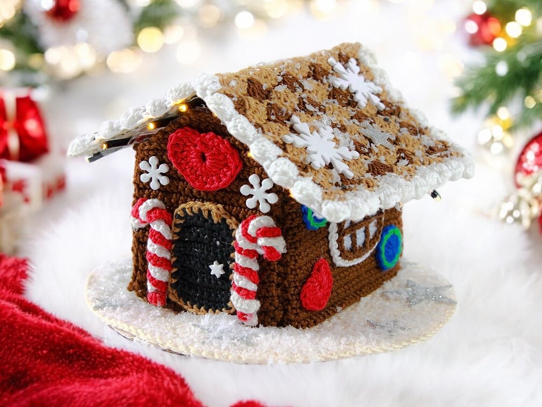 Small crocheted gingerbread house with scalloped white roof, candy-cane pillars, snowflake appliqués and red heart on a faux-snow surface