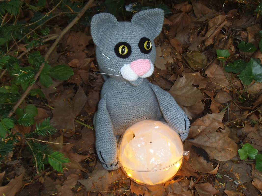 Gehäkelte graue Katze mit gelben Augen, rosa Nase und weißer Schnauze hält eine leuchtende Kugel auf Herbstlaub.