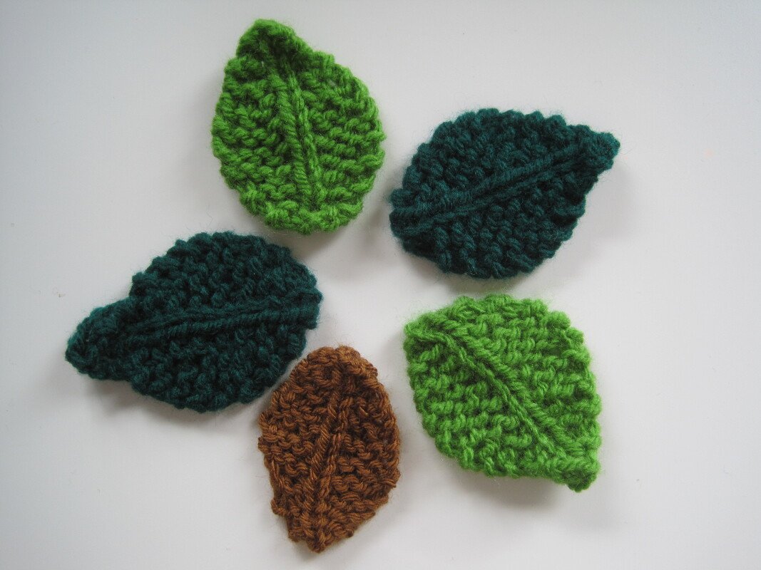 Fünf gestrickte Blatt-Applikationen in verschiedenen Grüntönen und Braun auf weißem Hintergrund