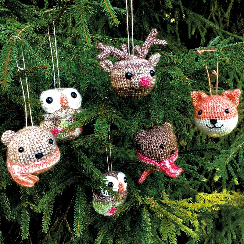 Sechs gehäkelte Tier-Weihnachtsanhänger (Rentier, zwei Eulen, zwei Bären, Fuchs) an grünen Tannenzweigen.