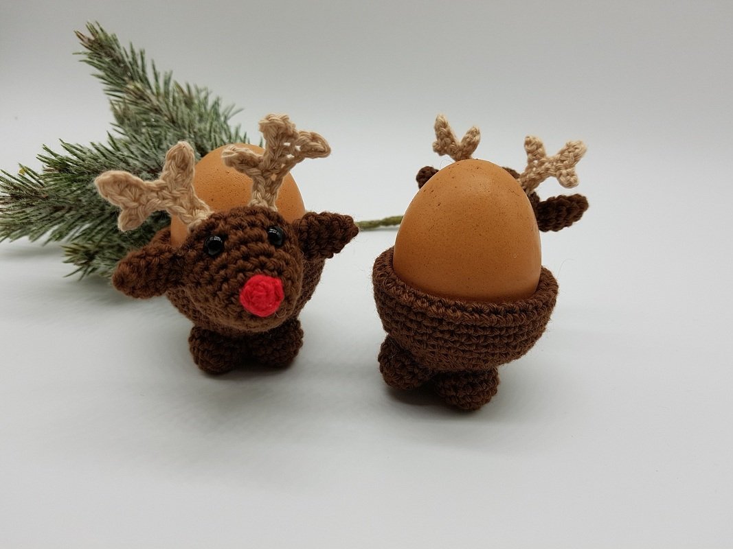 Zwei braune gehäkelte Rentier-Eierwärmer mit Geweih, eines mit roter Nase, jeweils mit Ei vor einem Tannenzweig.