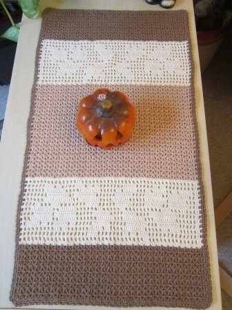 Gehäkelter Tischläufer in Dunkelbraun, Hellbraun und Creme mit durchbrochenem Herzmuster, auf einem Tisch mit Kürbisdekoration.