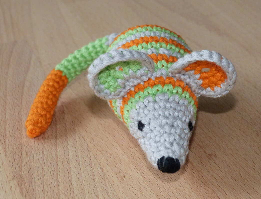 Gehäkelte Babyrassel in Form einer Maus mit orange-grünen Streifen und schwarzer Nase auf hellem Holz
