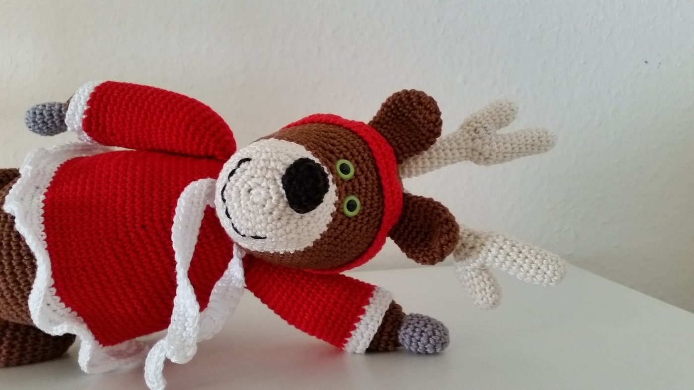 Gehäkeltes Rentier-Amigurumi in rotem Nikolaus-Kostüm mit weißen Akzenten, grünen Augen und beigem Geweih auf hellem Hintergrund.