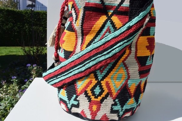 Gehäkelte Tasche mit geometrischem Ethno-Muster in Rot, Gelb, Türkis und Schwarz auf weißer Fläche im Freien.