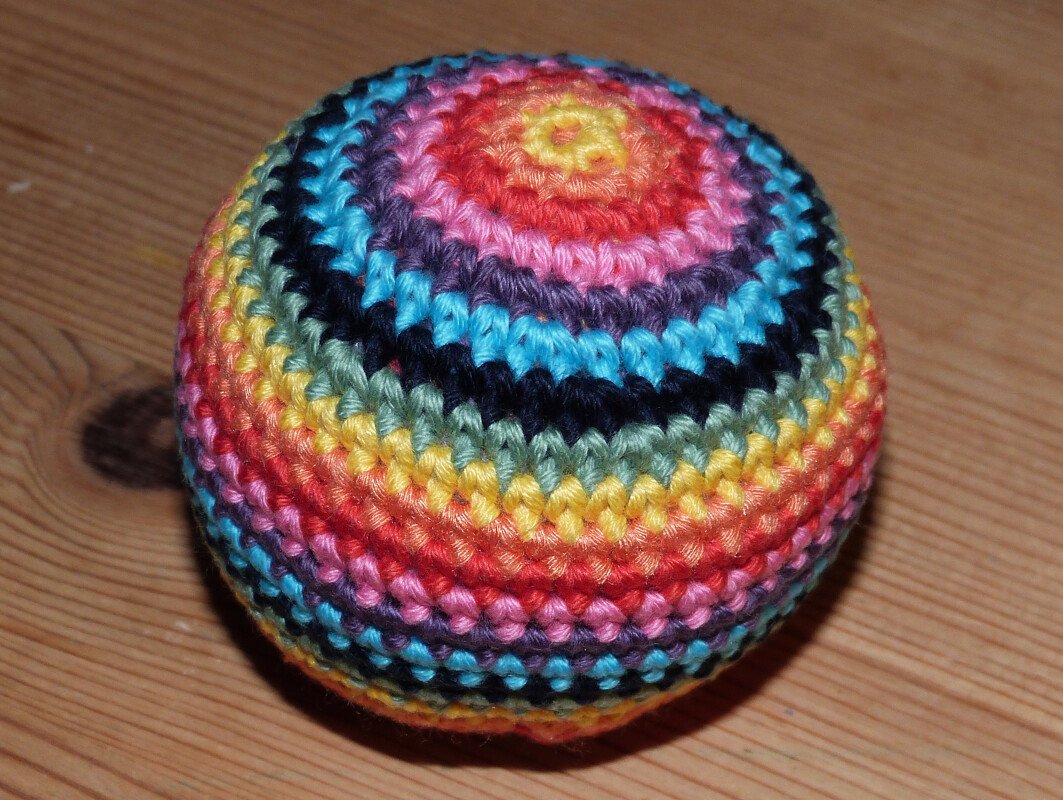 Runder gehäkelter Spielball mit bunten Streifen in Regenbogenfarben auf Holzuntergrund.