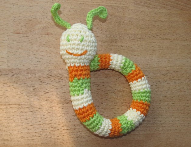 Gehäkelter Rasselring in Orange, Grün und Weiß mit kleinem Käferkopf und Antennen auf Holzuntergrund
