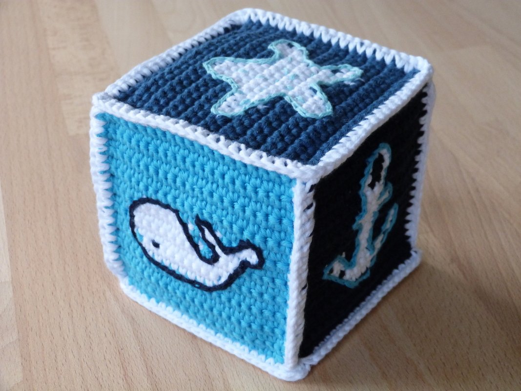 Gehäkelter Spielwürfel in Blau und Weiß mit applizierten Seestern-, Wal- und Anker-Motiven auf hellem Holztisch.