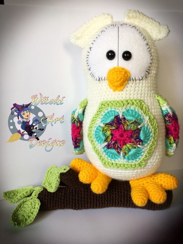 Amigurumi-Eule in Creme mit mehrfarbigem Häkelmandala auf Brust und bunten Flügeln auf einem gehäkelten Ast.