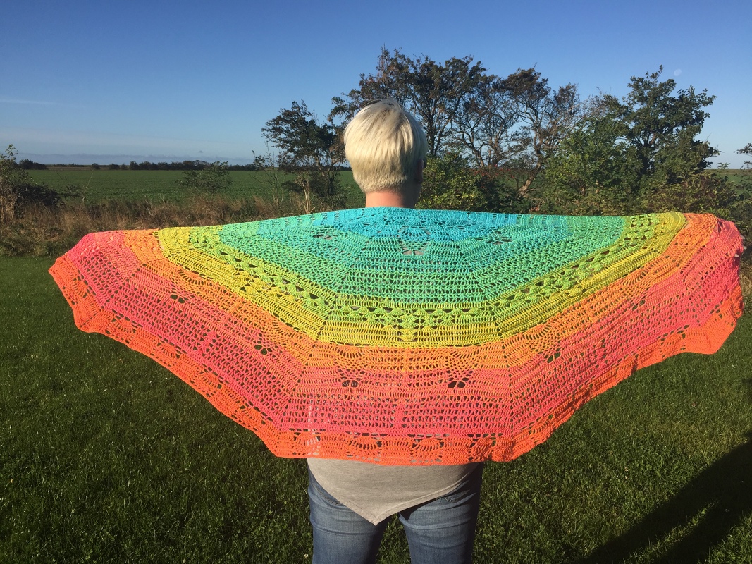 Person von hinten trägt ein weit ausgebreitetes halbrundes Häkel­tuch mit Farbverlauf von Türkis bis Pink vor einer ländlichen Kulisse.