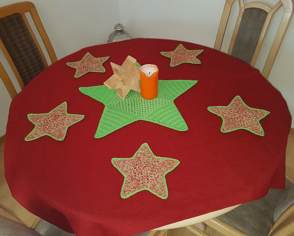 Runder Tisch mit roter Decke, großem grünen Häkelstern in der Mitte, sechs gehäkelten Stern-Untersetzern, Holzstern und orangefarbener Kerze