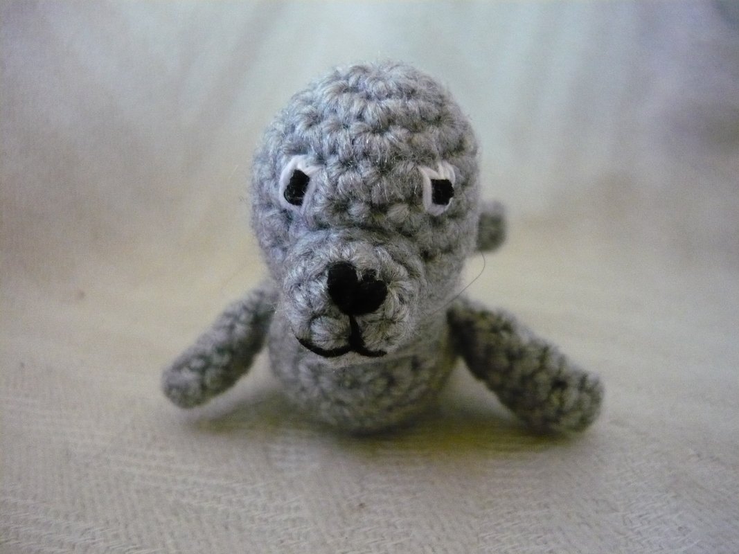 Gehäkelte graue Amigurumi-Robbe mit gestickter Nase und Augen auf hellem Hintergrund