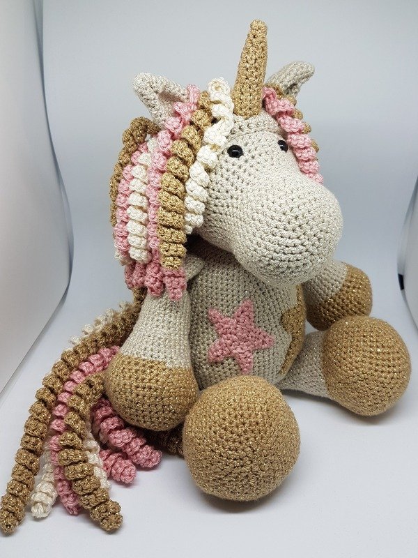 Gehäkeltes Amigurumi-Einhorn in Beige mit goldenem Horn und lockiger Mähne in Rosa, Creme und Gold, sitzend