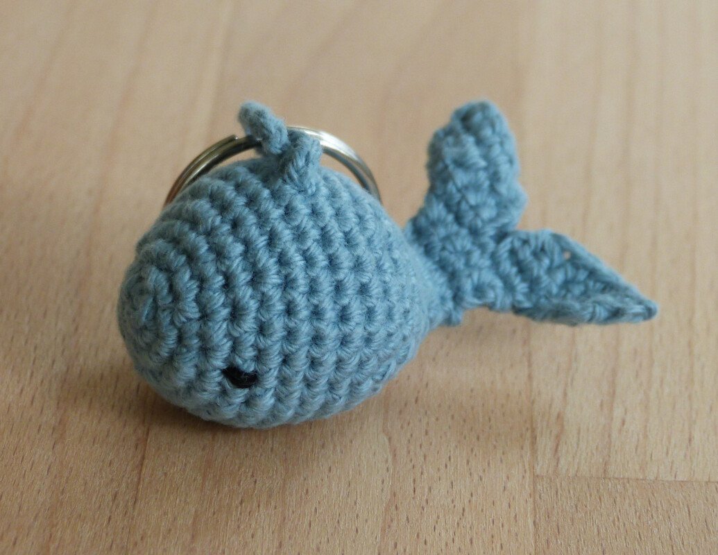 Gehäkelter hellblauer Wal mit Metall-Schlüsselring auf heller Holzoberfläche, sichtbare Häkelmaschen und kleines schwarzes Auge