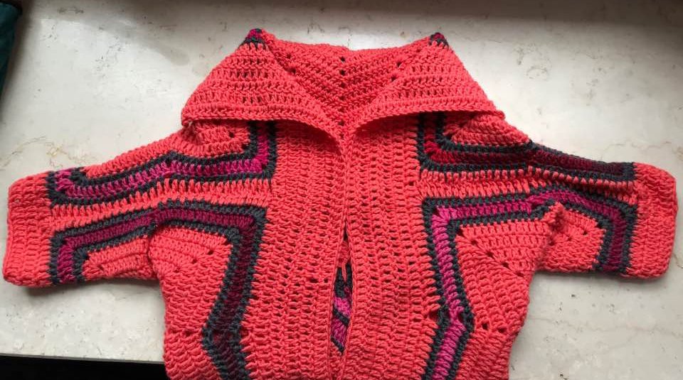 Gehäkelte Damenjacke in Korallenrot mit breitem Kragen und Streifen in Dunkelgrau und Fuchsia, flach liegend fotografiert.