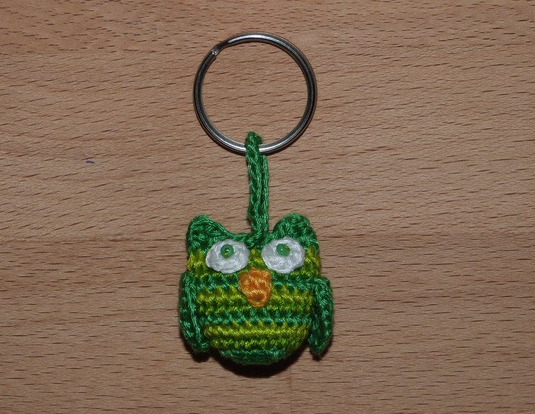 Gehäkelter grün-gelb gestreifter Eulen-Schlüsselanhänger mit weißen Augen und orangem Schnabel an Metallring auf Holzuntergrund