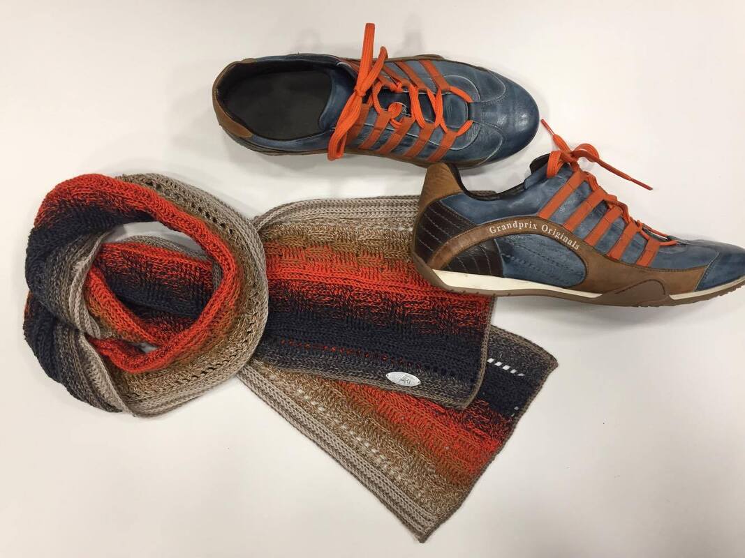 Gehäkelter Farbverlaufsschal in Orange, Blau und Beige neben zwei blauen Freizeitschuhen auf hellem Untergrund.