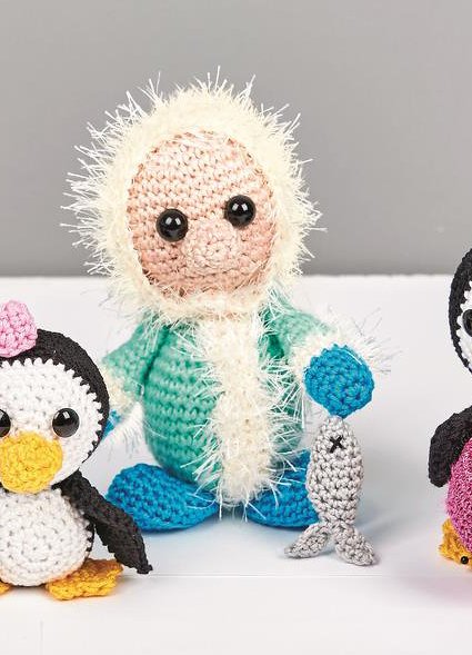 Gehäkelte Figur in türkisem Winteroutfit mit flauschiger Kapuze hält einen kleinen grauen Fisch; links und rechts zwei gehäkelte Pinguine auf weißem Untergrund.