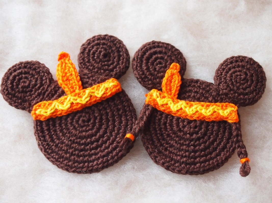 Two brown crocheted appliques with round ears and orange and yellow headbands; the right applique has a short braided tail.