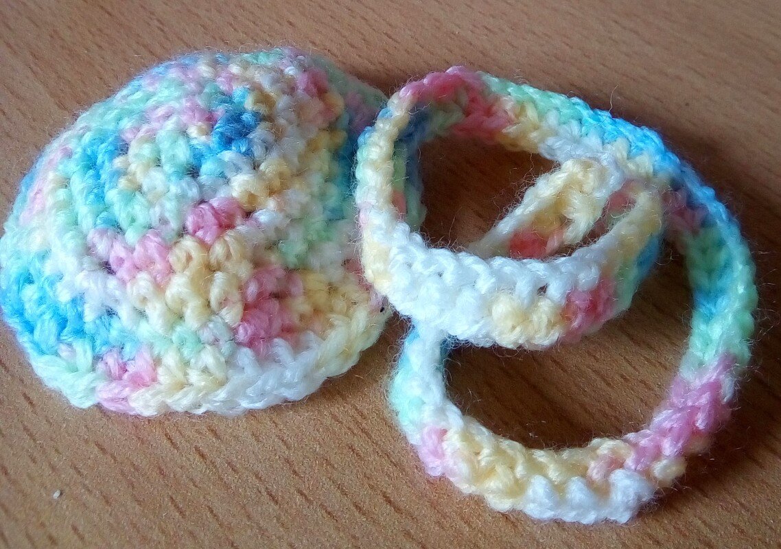 Ein flaches, rundes Häkelstück und ein geformter Häkelring in sanften Pastellfarben auf einer Holzoberfläche