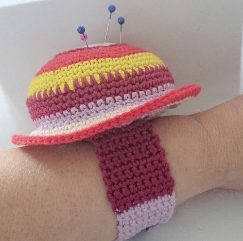 Gehäkeltes, gestreiftes Armnadelkissen in Rot, Gelb, Rosa und Flieder mit Stecknadeln, am Handgelenk befestigt.