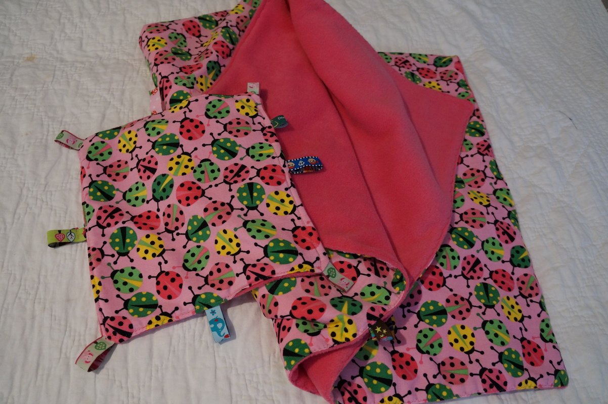 Rosa Babydecke mit Marienkäfer-Muster und kleines Tasttuch mit bunten Stofflaschen auf weißer Unterlage