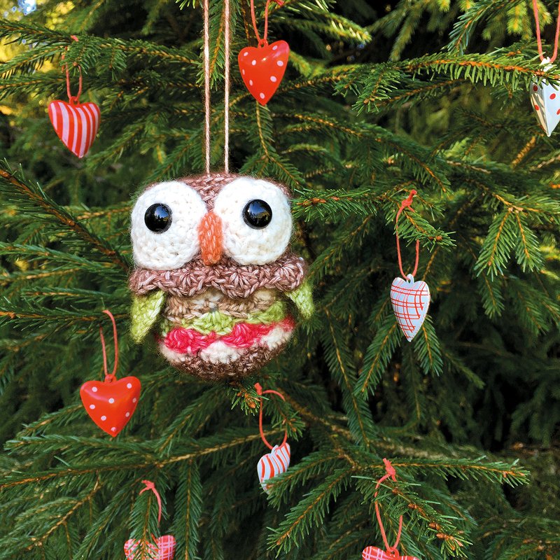 Gehäkelte Eule mit großen Augen als Weihnachtsbaum-Anhänger an einem Tannenzweig, umgeben von kleinen Herzanhängern