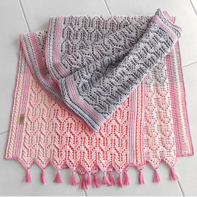 Gefaltete gehäkelte Decke in Rosa und Grau mit Ajourmuster und Quasten am Saum, auf hellem Boden liegend.