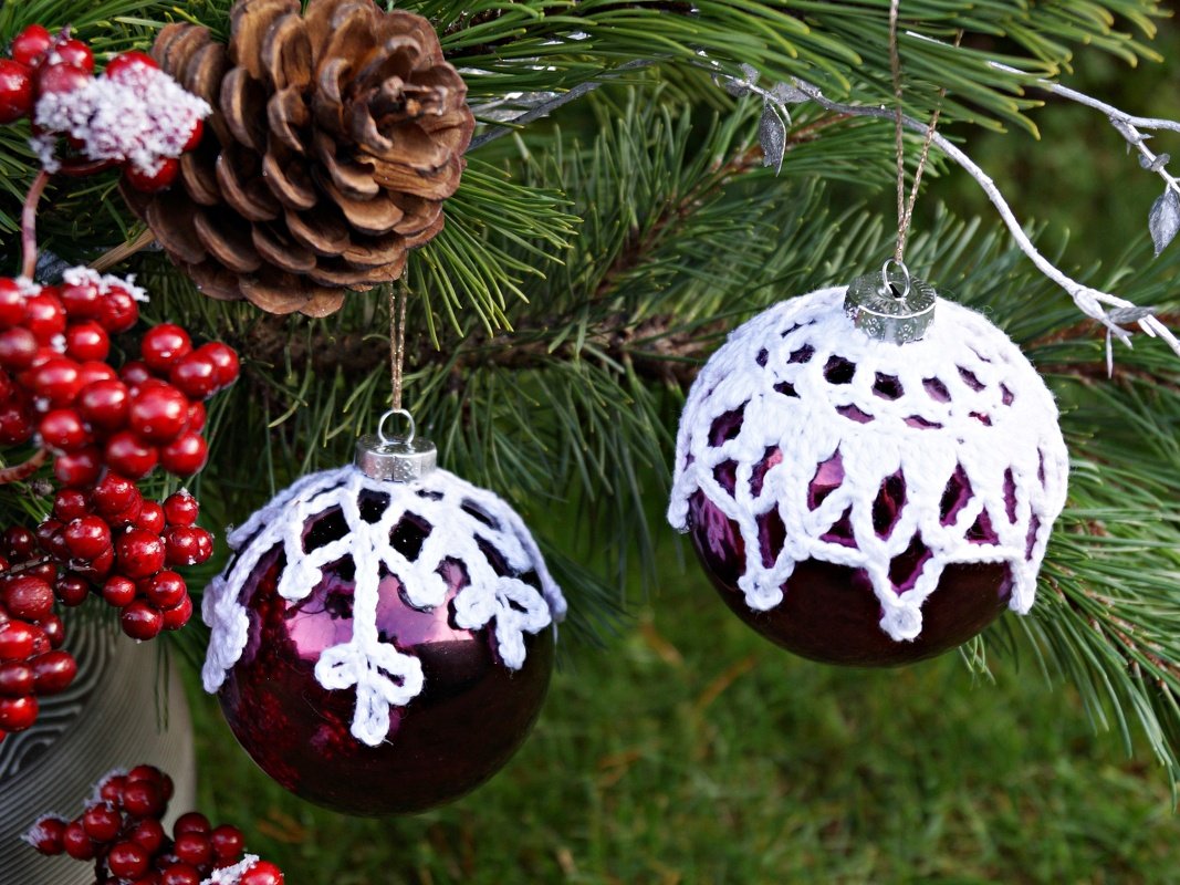Zwei violette Christbaumkugeln mit weißen gehäkelten Häubchen an einem Kiefernzweig