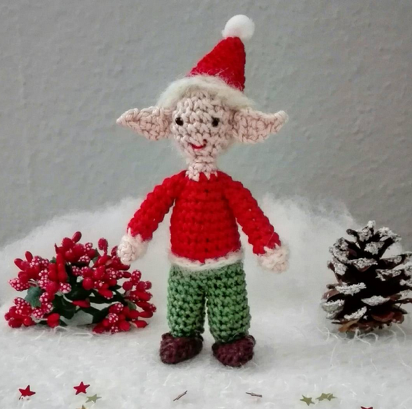 Gehäkelter Weihnachtswichtel mit roter Zipfelmütze, rotem Oberteil und grüner Hose vor weihnachtlicher Dekoration