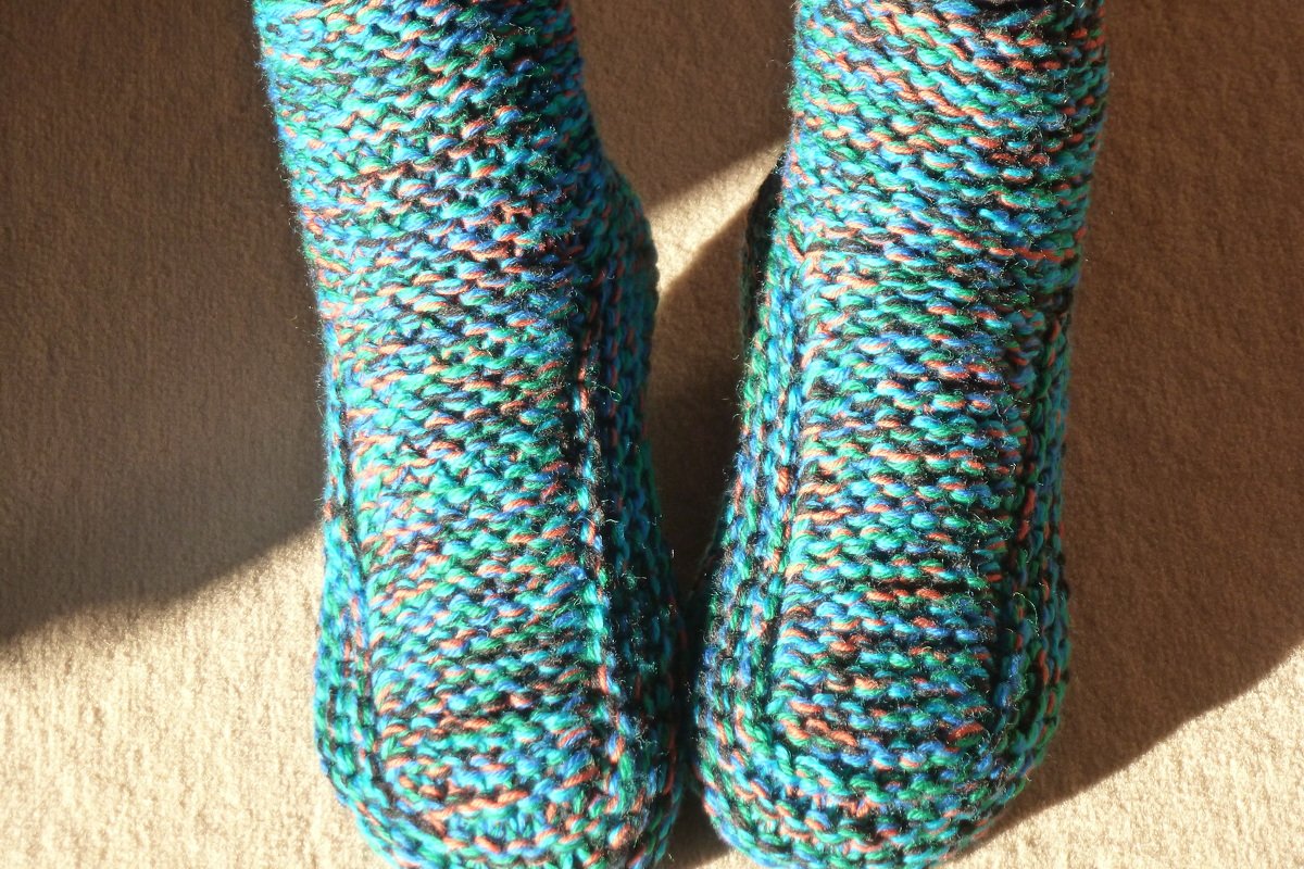 Nahaufnahme eines Paars gestrickter mehrfarbiger Hausschuhe (türkis, grün, orange) auf beigem Teppich im Sonnenlicht.