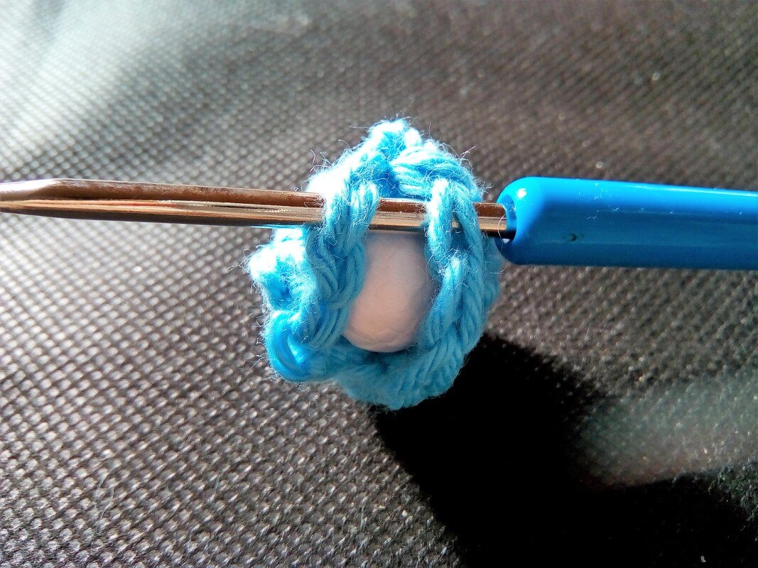 Blaue gehäkelte Maschen um eine kleine weiße Kugel auf einer Häkelnadel