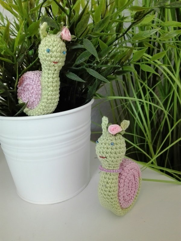 Zwei gehäkelte Amigurumi‑Schnecken in Hellgrün mit rosa Häuschen und kleinen rosa Rosen, eine im weißen Blumentopf, die andere daneben vor grünen Pflanzen.