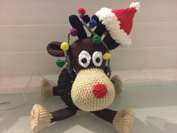Handgehäkeltes Rentier mit braunem Körper, beiger Schnauze, roter Nase, weißen Augen, roter Weihnachtsmütze und bunter Lichterkette um das Geweih.
