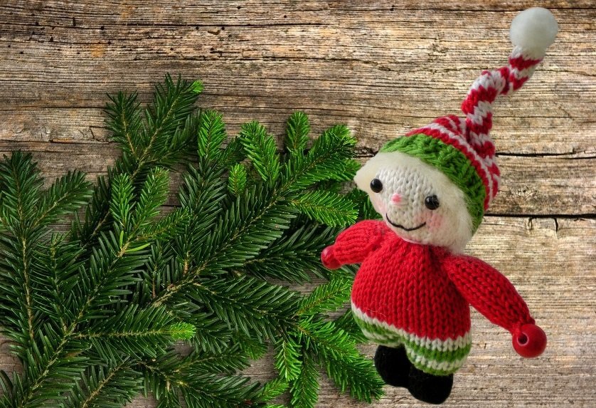 Gestrickte Amigurumi-Figur in Rot und Grün mit rot-weiß gestreifter Zipfelmütze neben Tannenzweigen auf Holzhintergrund