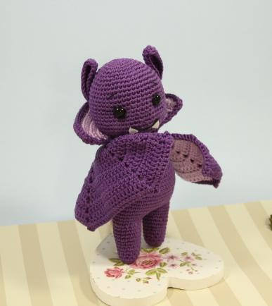 Purple crocheted bat amigurumi with wings and small fangs standing on a heart-shaped floral base