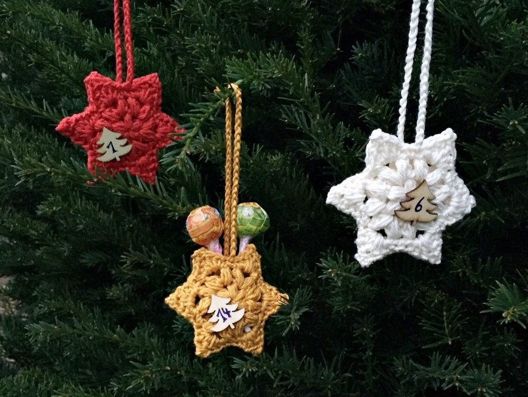 Drei kleine gehäkelte Stern-Taschen mit Schlaufen und nummerierten Holzplättchen, an einem Weihnachtsbaum aufgehängt; eine Tasche enthält Lollis