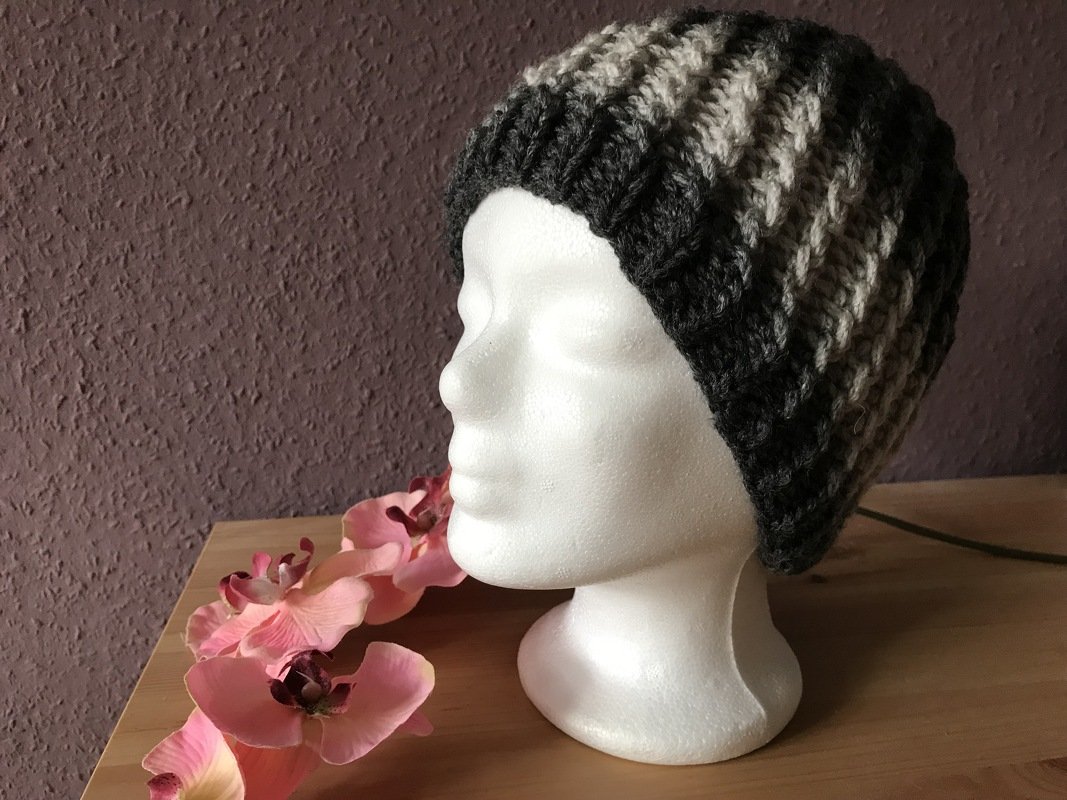 Gestrickte gestreifte Mütze in Dunkel- und Hellgrau auf weißem Schaufensterkopf, rosa Kunstorchideen daneben
