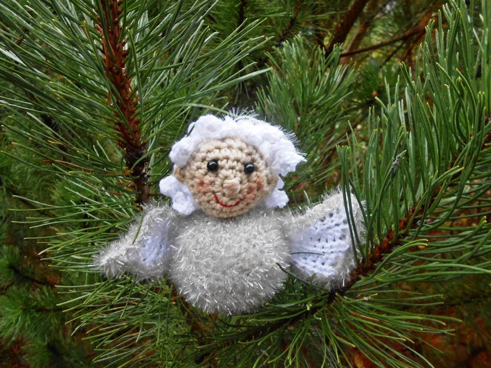 Silberweißes gehäkeltes Amigurumi-Engelchen mit flauschigem Körper und weißen Flügeln an einem Kiefernzweig
