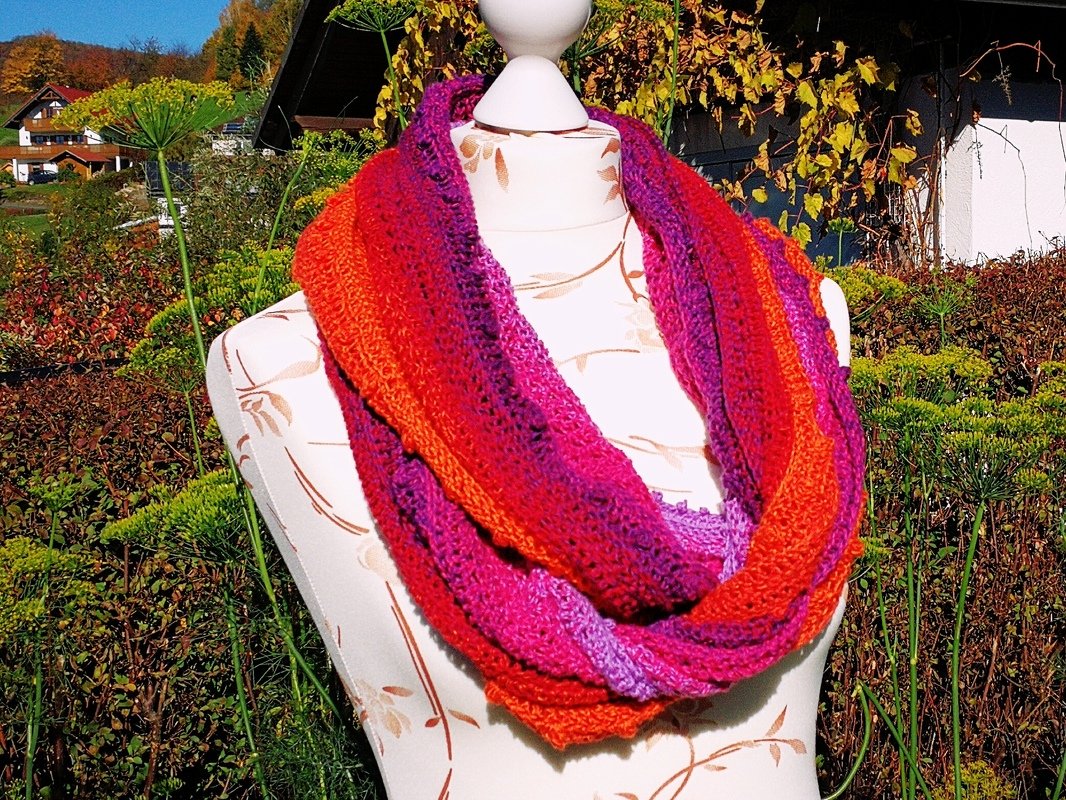 Gehäkelter Loop‑Schal in Orange, Rot und Lila auf einer Puppe vor herbstlicher Gartenkulisse.