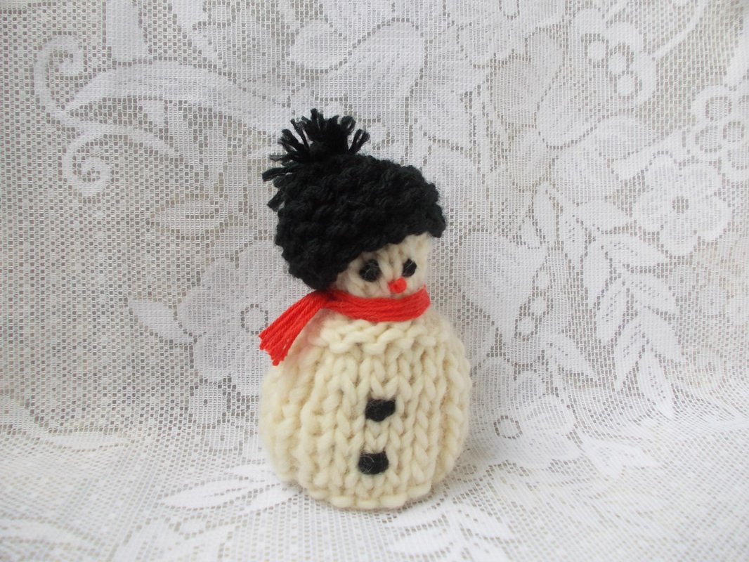 Gestrickter Mini‑Schneemann in Creme mit schwarzer Bommelmütze und rotem Schal vor Spitzenstoff
