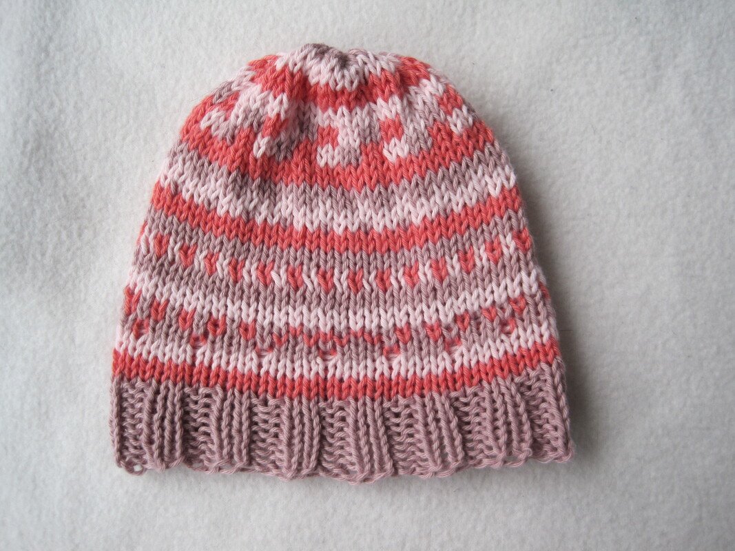Nahaufnahme einer gestrickten Babymütze mit Herz- und Streifenmuster in Rosa, Koralle und Mauve auf hellem Hintergrund.