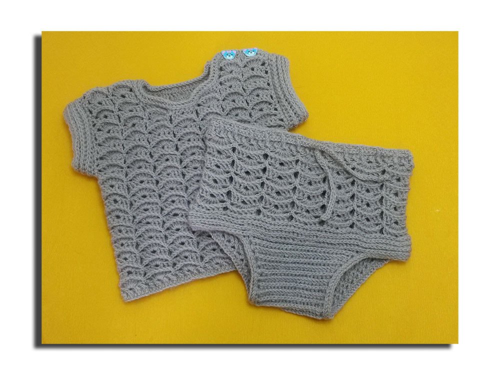 Grau gehäkeltes Baby-Set mit Muschelmuster, kurzärmeliges Oberteil mit zwei blauen Bärenknöpfen und passende kurze Hose auf gelbem Hintergrund.