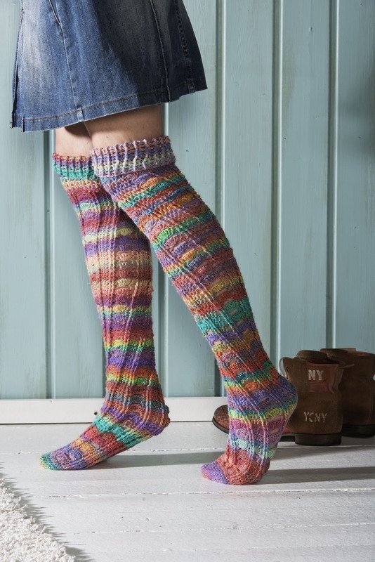 Beine in bunten, gehäkelten Overknee-Socken vor hellblauer Holzverkleidung.