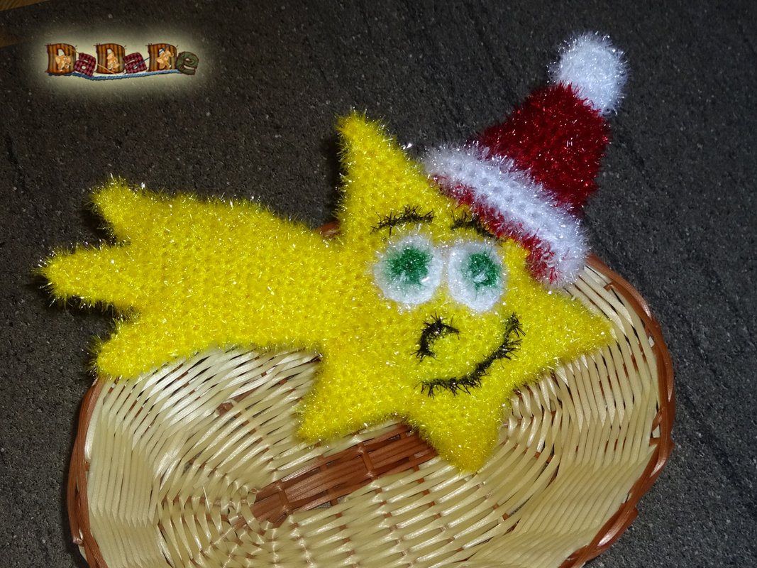 Gehäkelter gelber Stern mit grün-weißen Augen und roter Weihnachtsmütze auf geflochtenem Korb