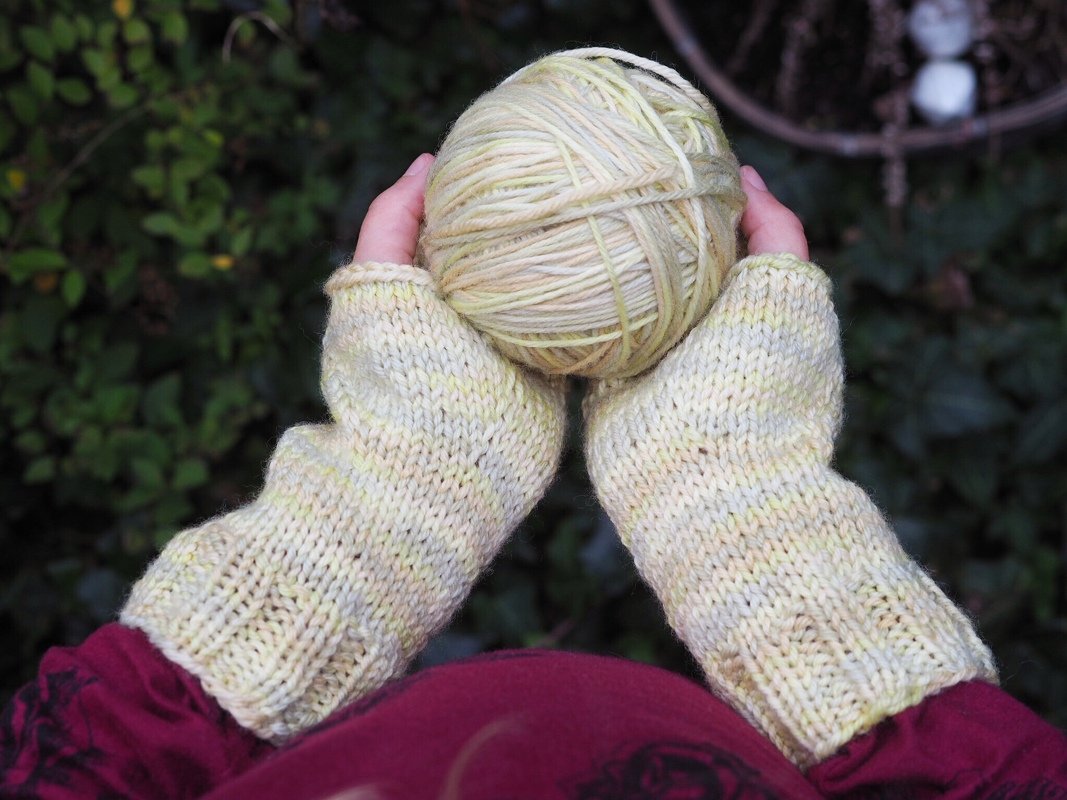 Hände mit gestrickten, fingerlosen Handschuhen in hellgelbem Garn halten ein Wollknäuel