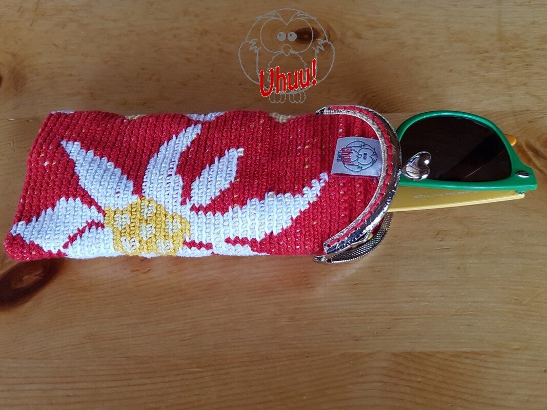 Gehäkeltes rotes Etui mit weiß-gelbem Edelweißmuster und Metallbügel, daneben eine grüne Sonnenbrille auf einem Holztisch.