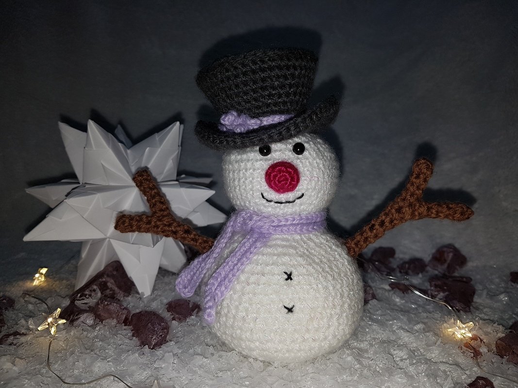 Gehäkelter Amigurumi‑Schneemann mit schwarzem Zylinder, lila Schal, roter Nase und Astarmen vor Papierstern und Lichterkette