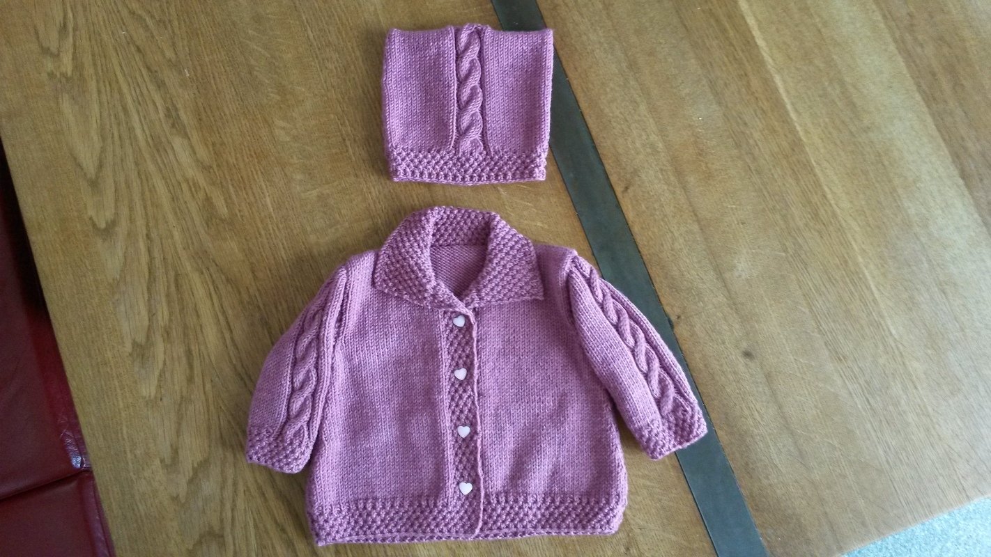 Mauvefarbenes gestricktes Baby-Set: Jacke mit Kragen und Herzknöpfen sowie passende Mütze mit Zopfdetail auf Holz.